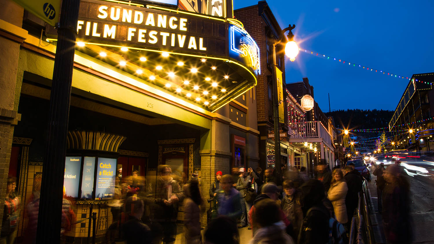 A busy Park City theater at night, with "Sundance Film Festival" illuminated on the sign, and lots of excited people in coats waiting under glowing lights