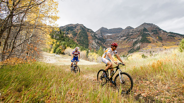 Two men on mountain bikes ride through golden Aspen leaves with breathtaking views of the Wasatch Mountains in the background