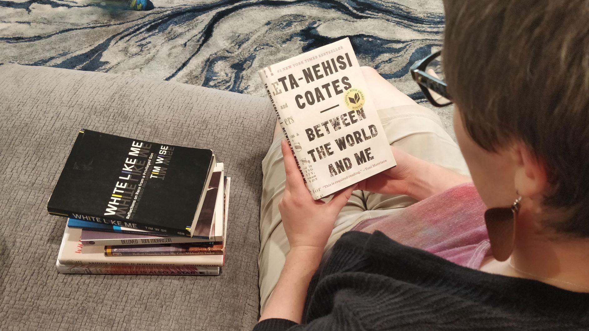 a woman reads between the world and me with a stack of more books beside her