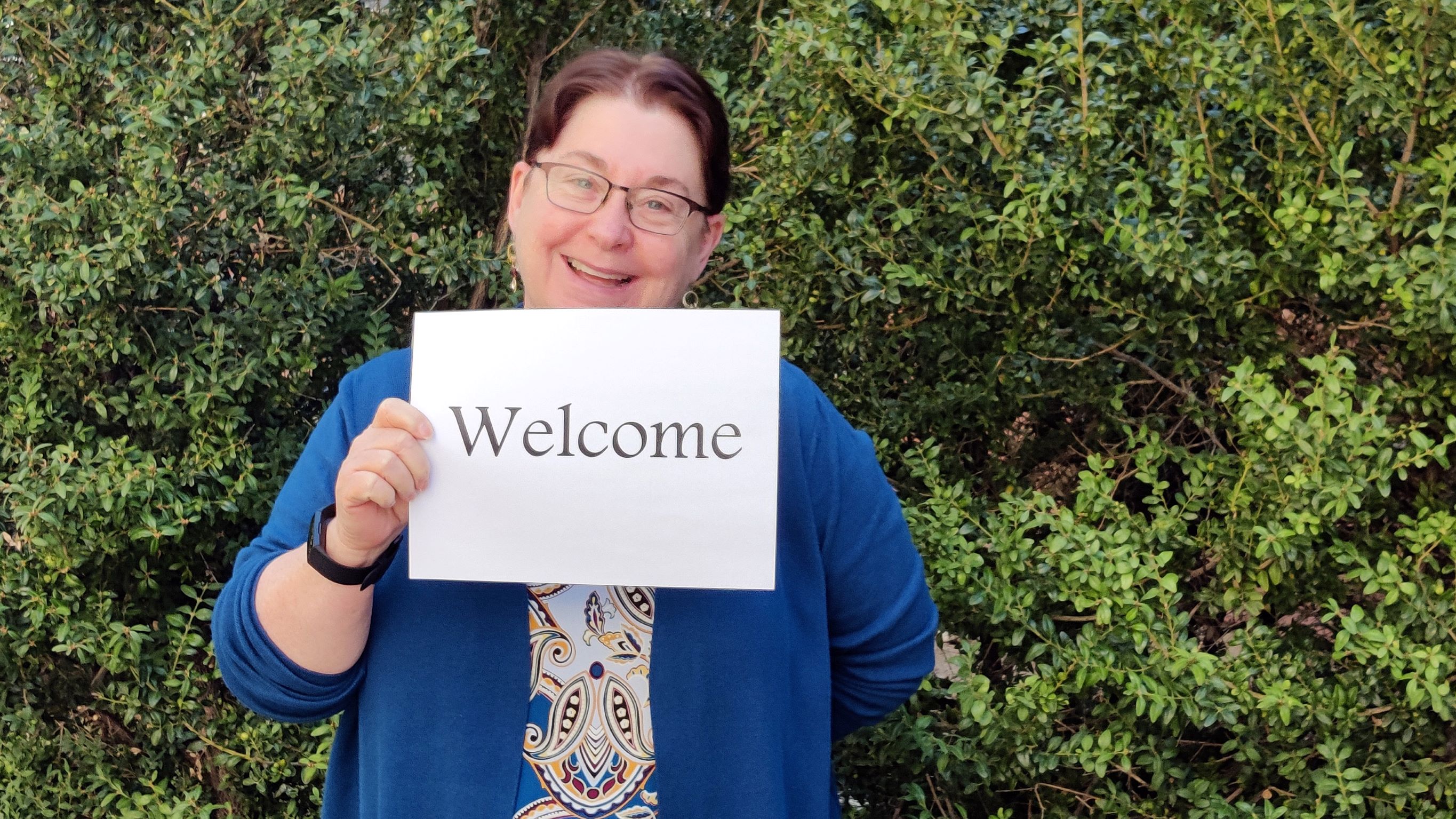 A middle aged white woman wearing glasses is smiling and holding a sign that says 