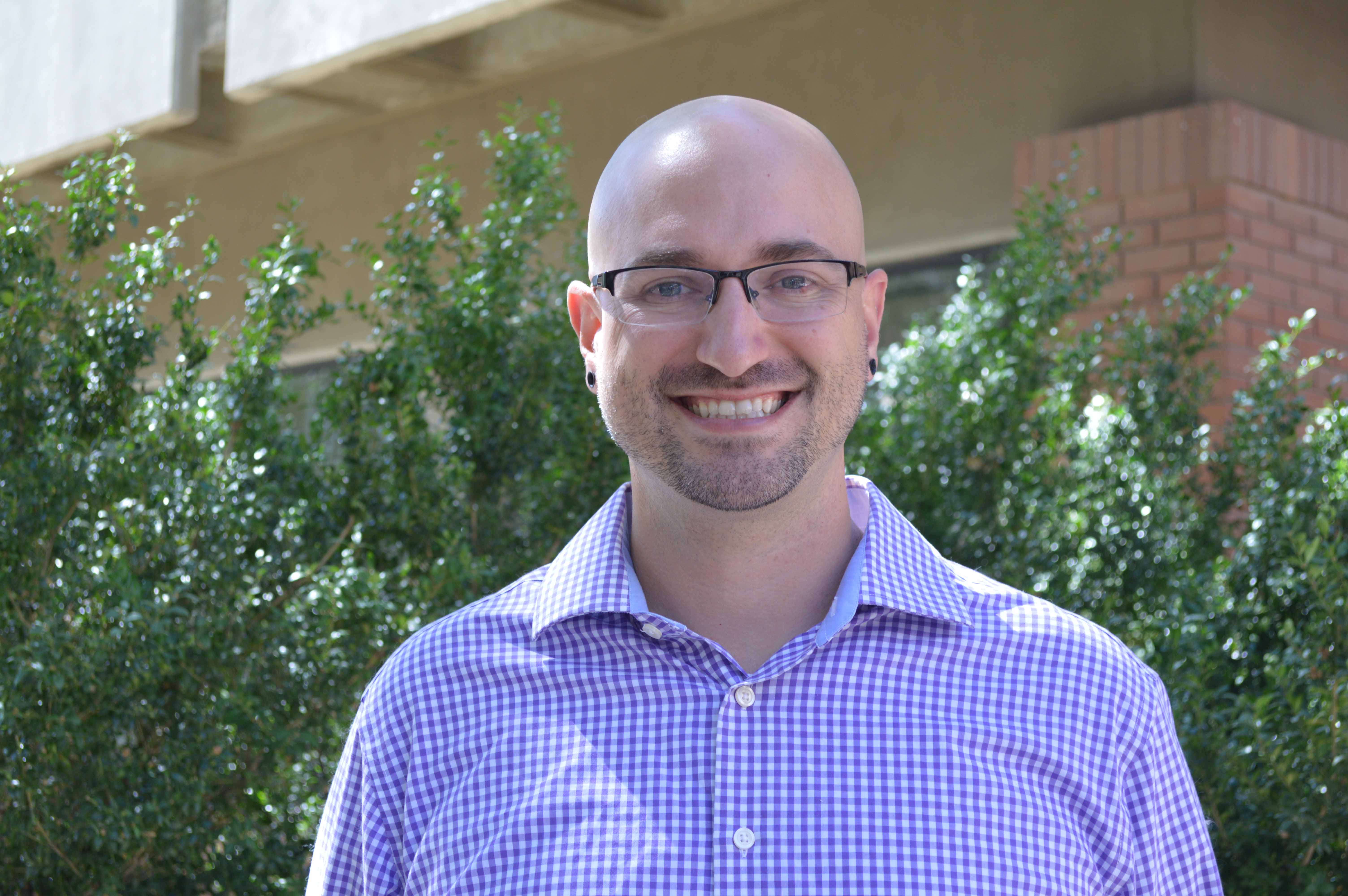 a middle aged white man wearing glasses smiles with trees and a brick building in the background