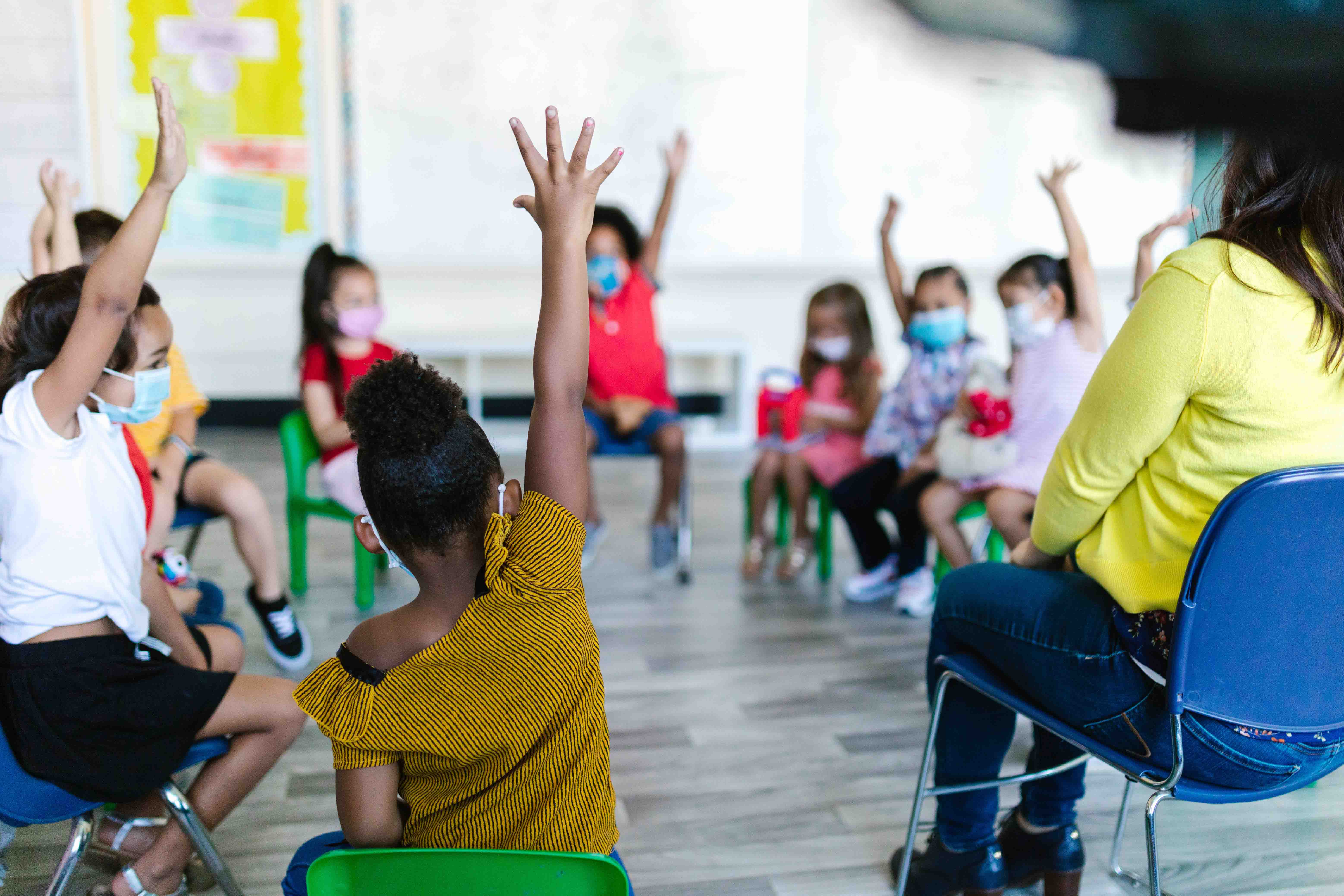 a group of masked children raise their hands as they sit in chairs in a circle