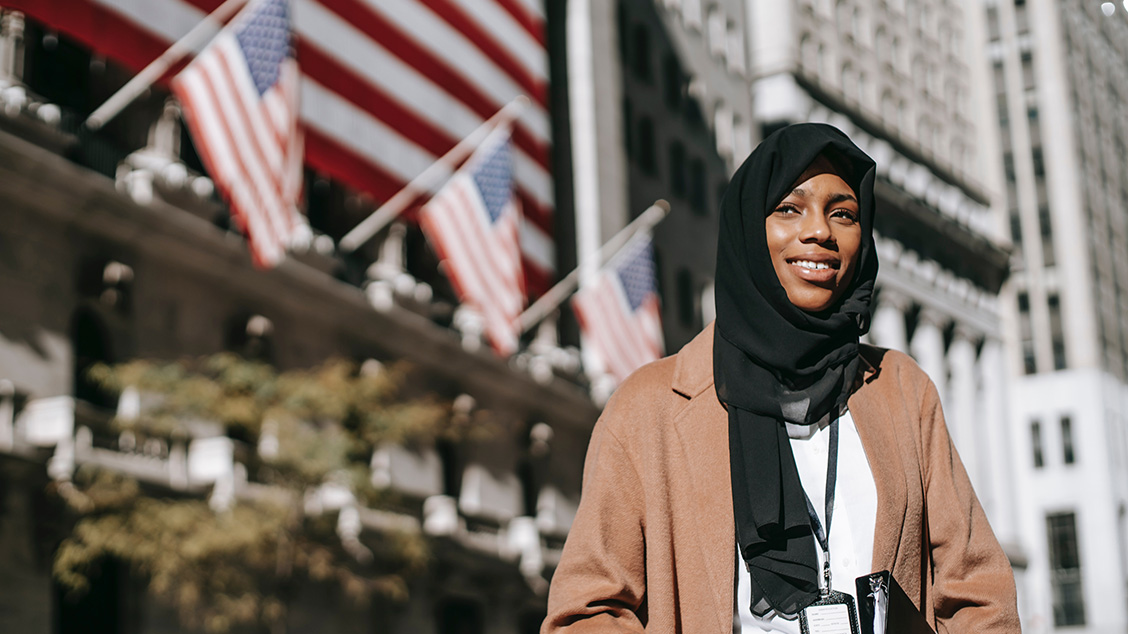 A woman of color wearing a black headscarf and a camel blazer smiles, looking away from the camera and holding a black binder, while standing in front of a tall building flying four United States flags.