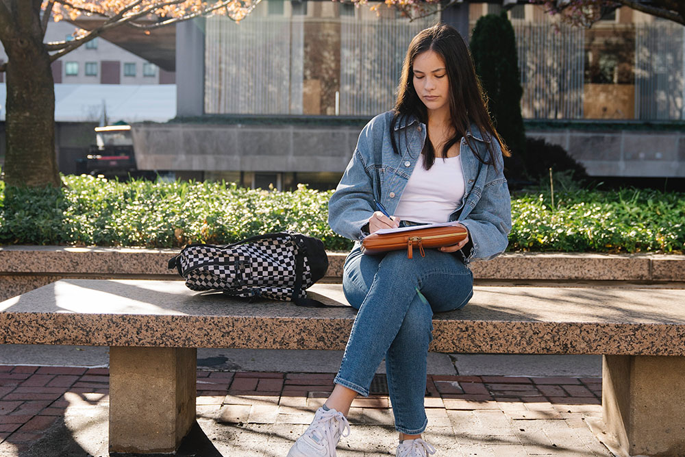 A female student sits on an outdoor bench, writing on a pad, a checkered backpack next to her