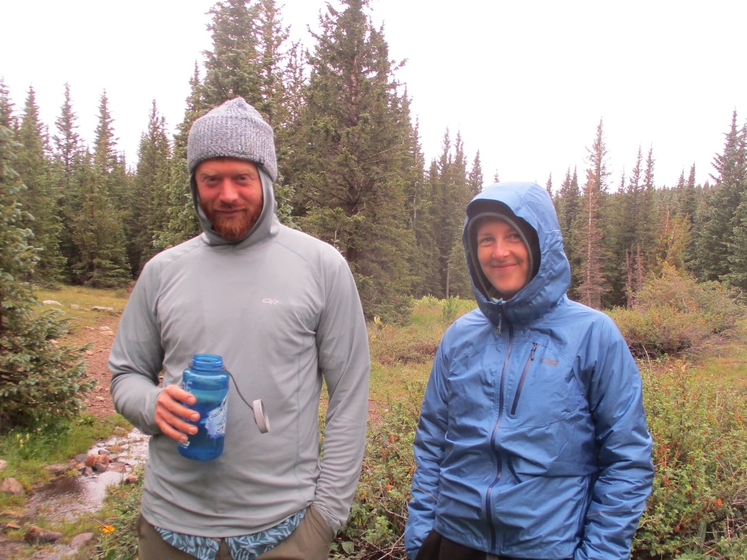 A man and a woman bundled up in coats, hats, and hoods. In the background the sky is gray and there are pine trees.