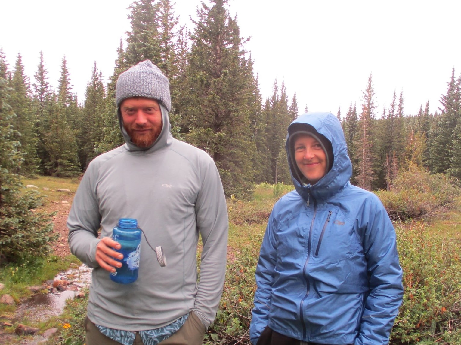 A man and a woman bundled up in coats, hats, and hoods.  In the background the sky is gray and there are pine trees.