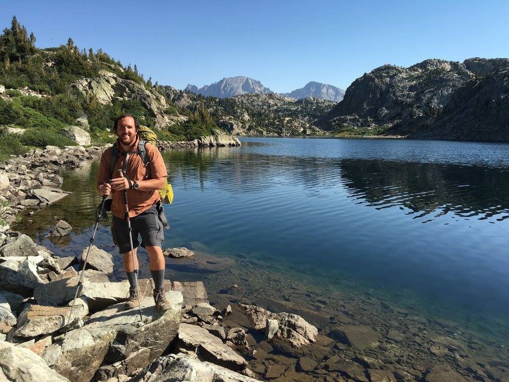 A man with walking sticks posing in front of a lake.  Mountains and trees rise up from the background, with rocks in the foregrounds