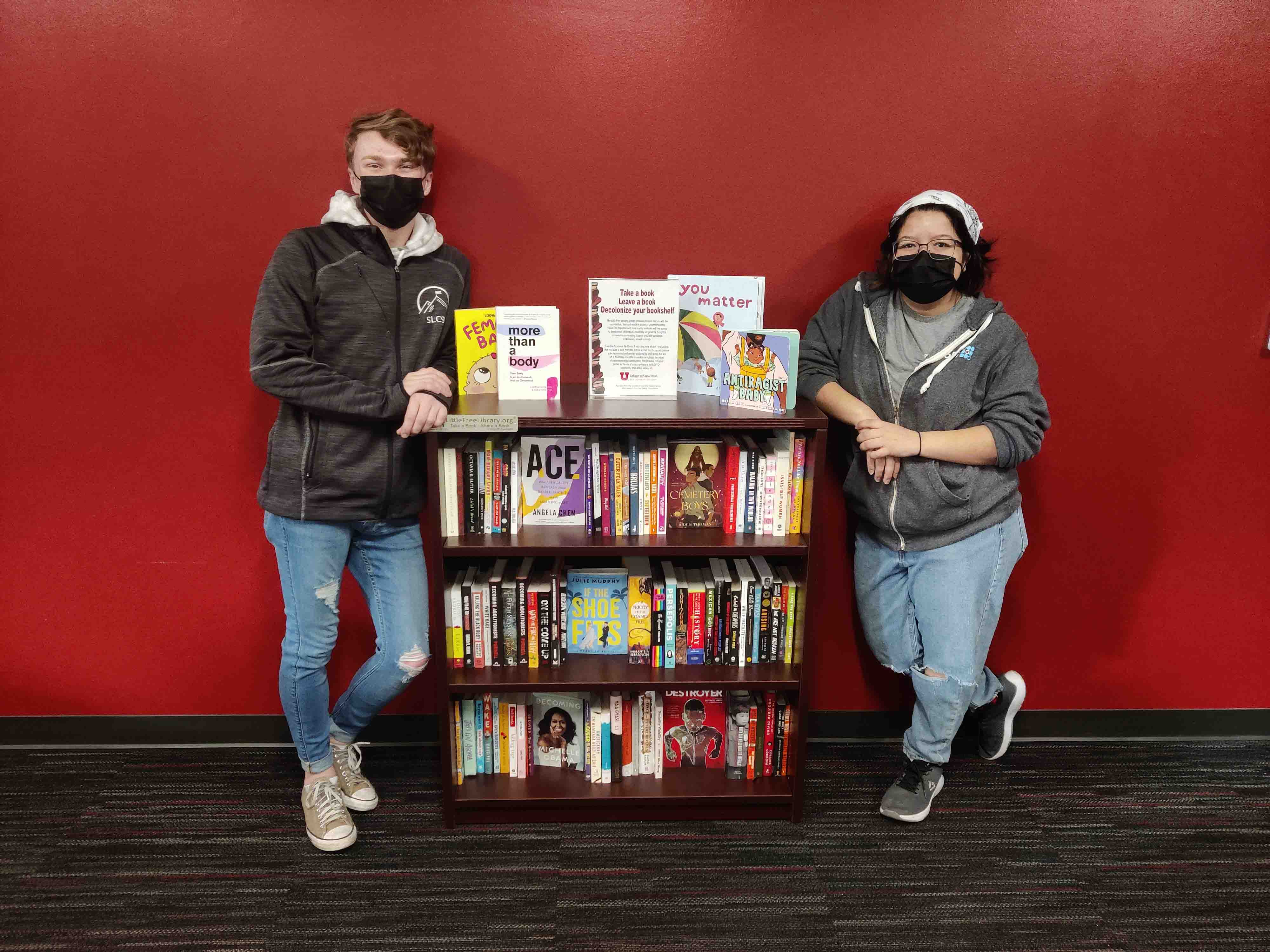 Two masked college students stand on opposite ends of a bookshelf full of books