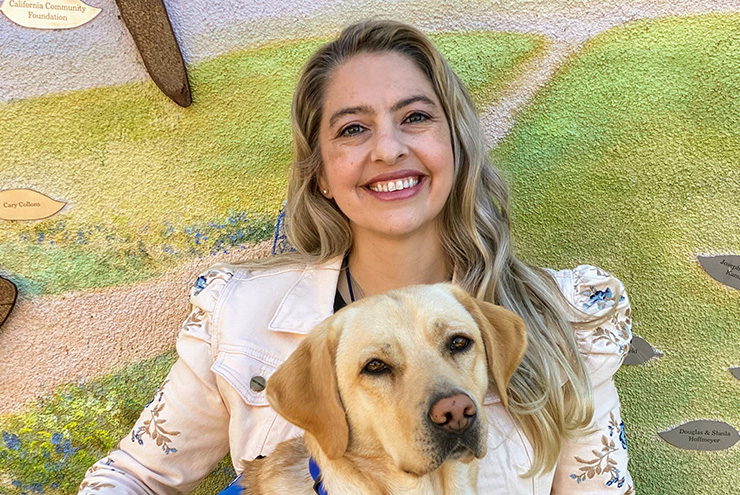 a white woman sits with a yellow lab therapy dog in a vest on her lab