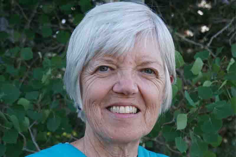 an older white woman with short gray hair smiles at the camera with green foliage in the background