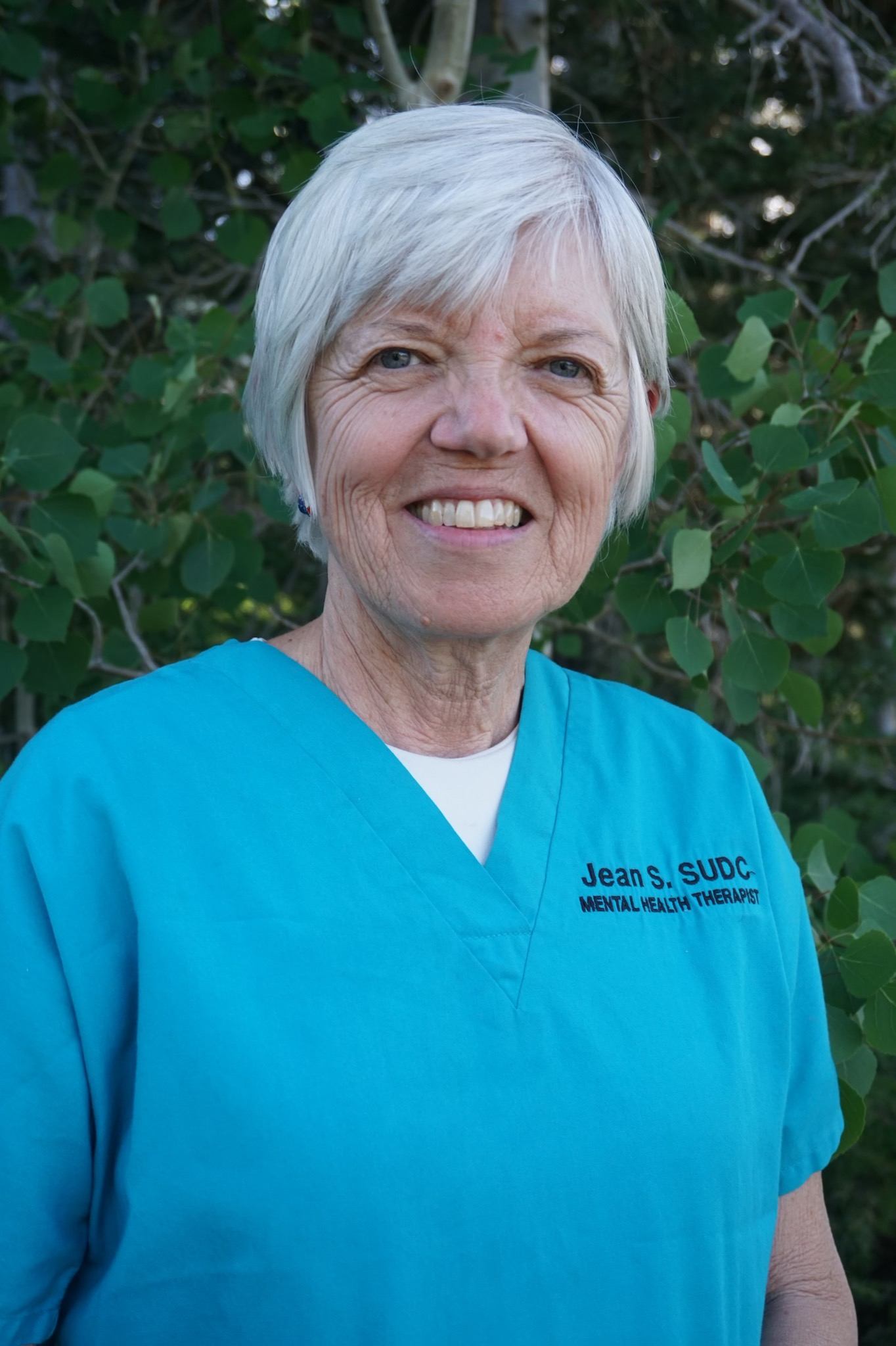 an older white woman with short gray hair smiles at the camera with green foliage in the background
