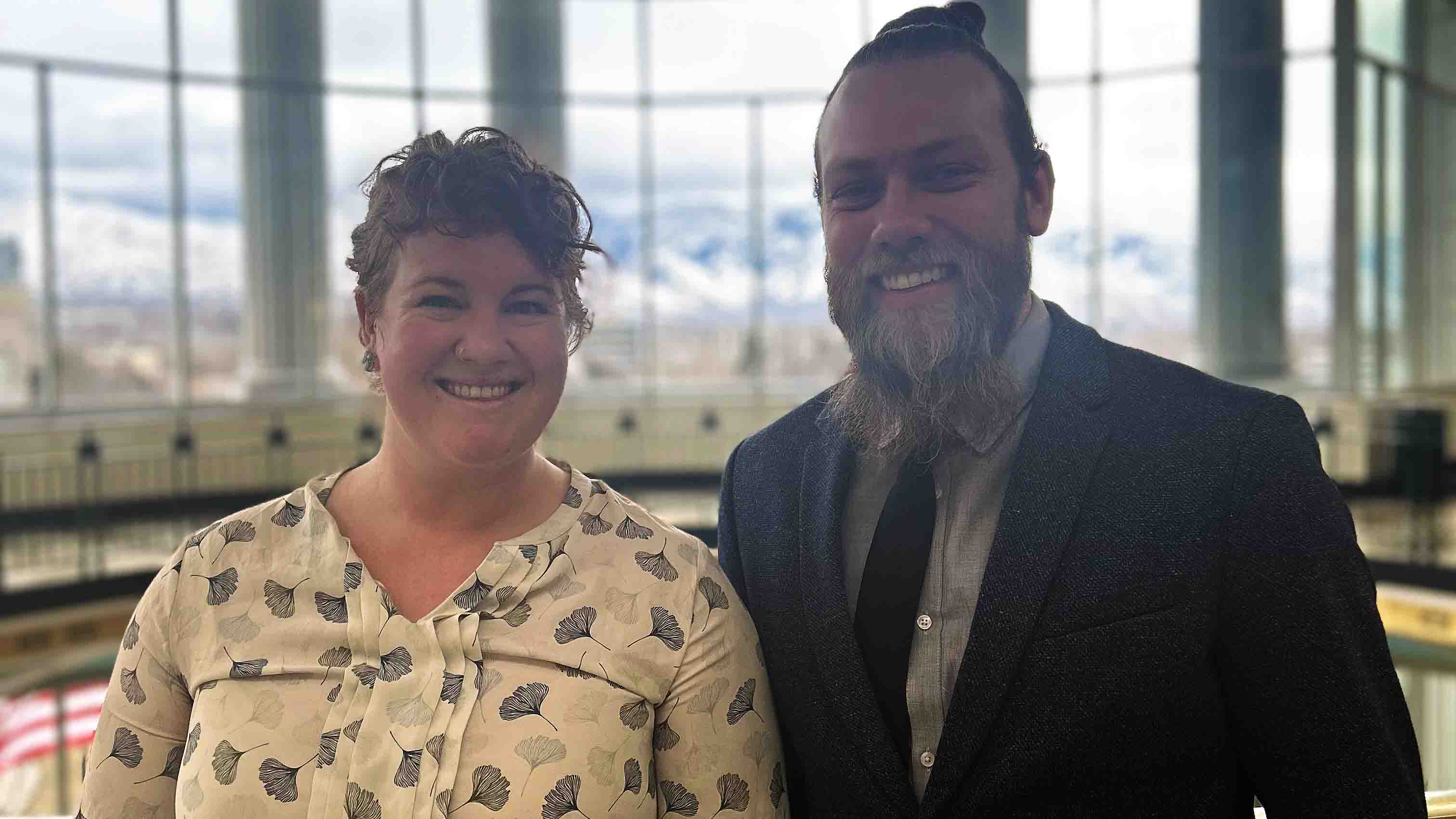 A white woman and white man looking into the camera and smile with windows in the background