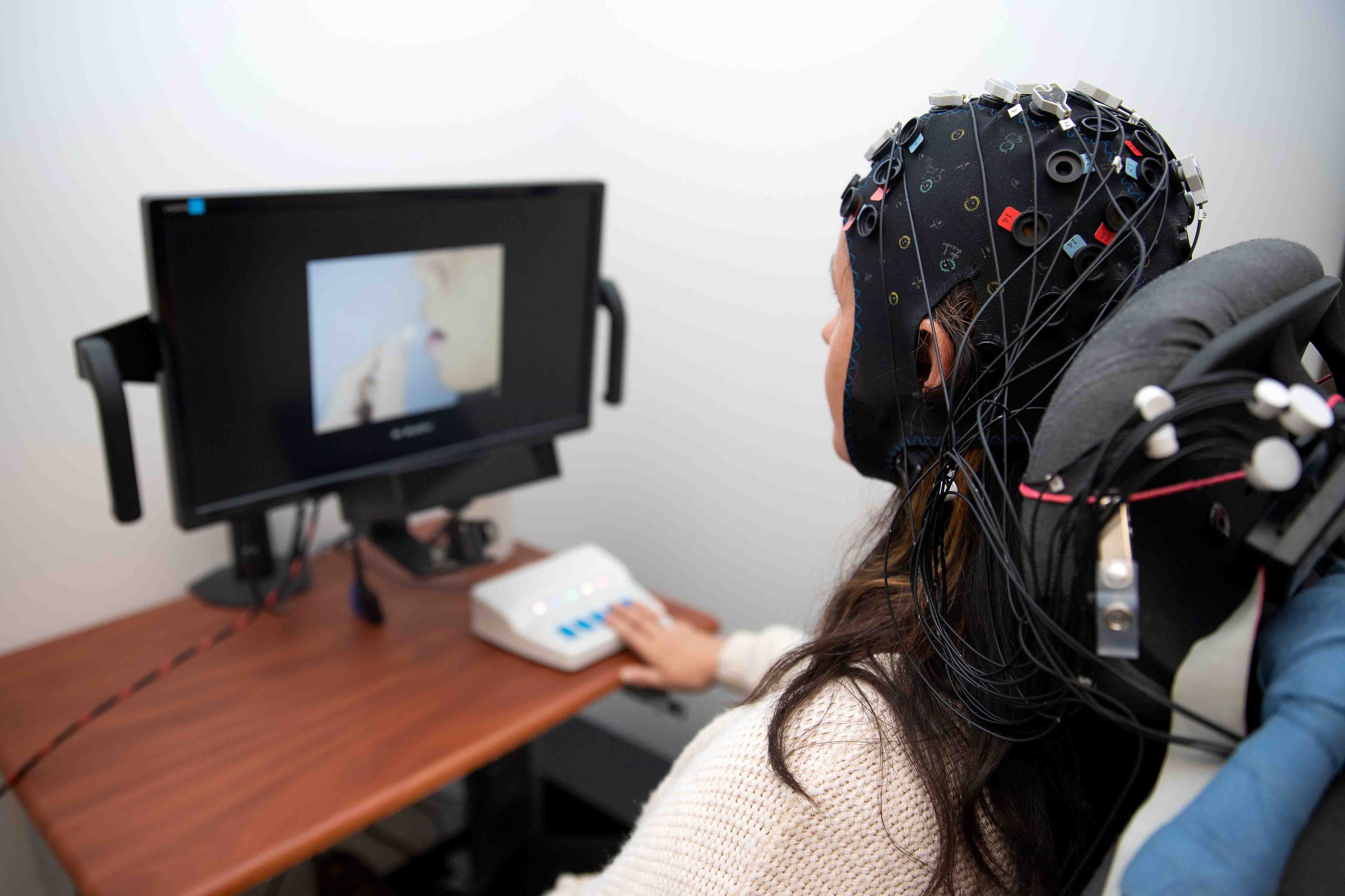 A woman sits in front of a computer screen. She wears a cap with many wires coming out of it.