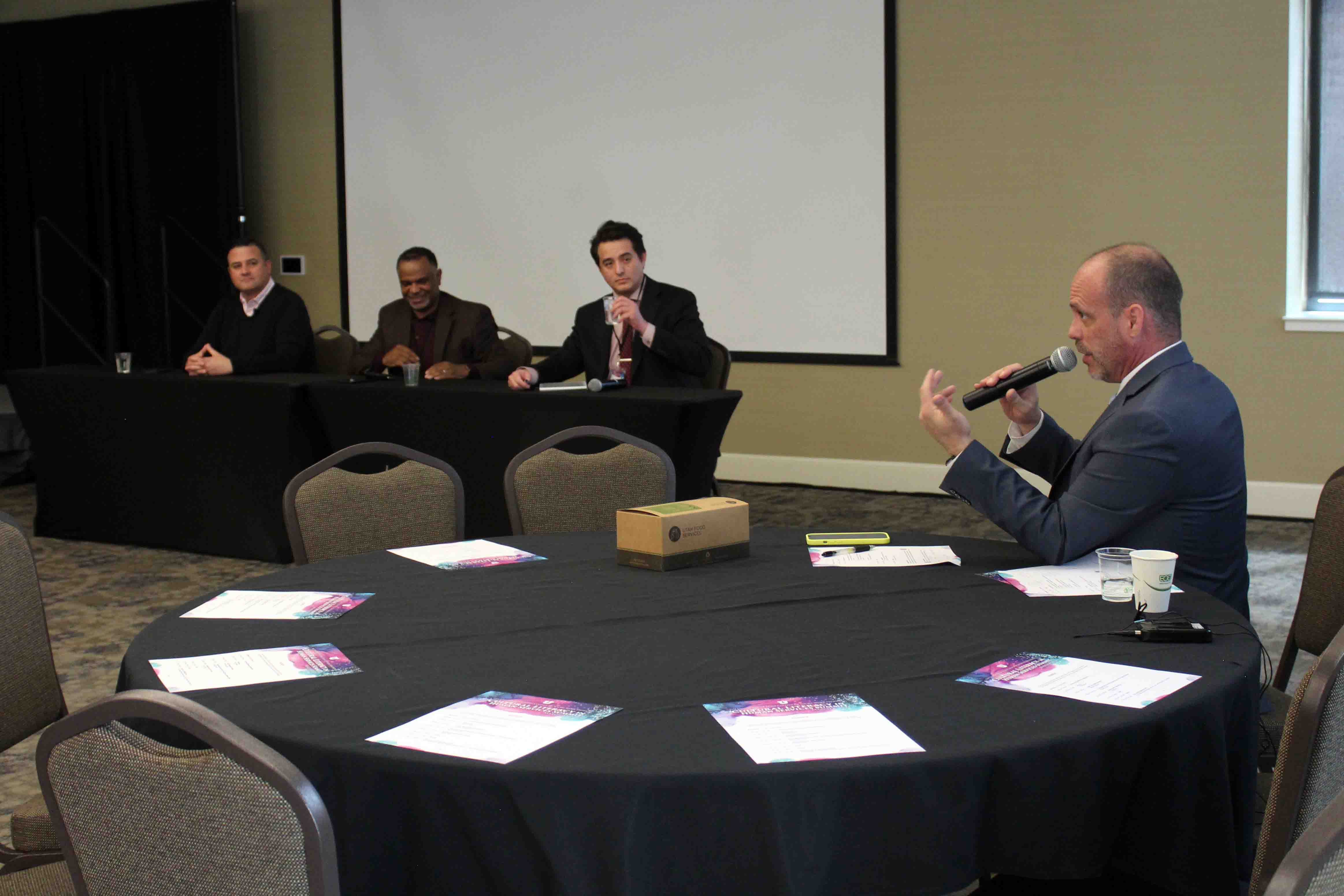 a white man sits at a round table speaking while others look at him from another table