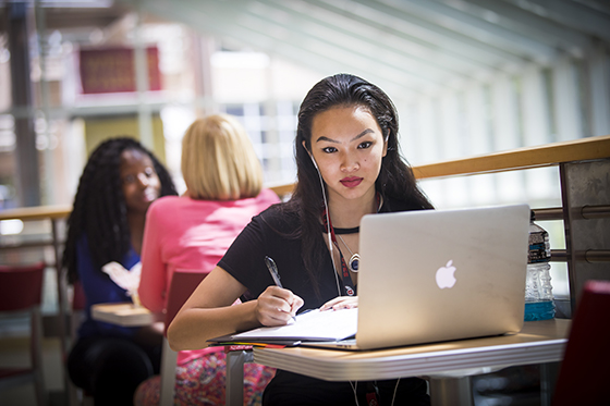 A young adult college student sits at a desk, looking at her laptop screen and writing in a notebook with headphones in her ears. . Other students sit in the background.