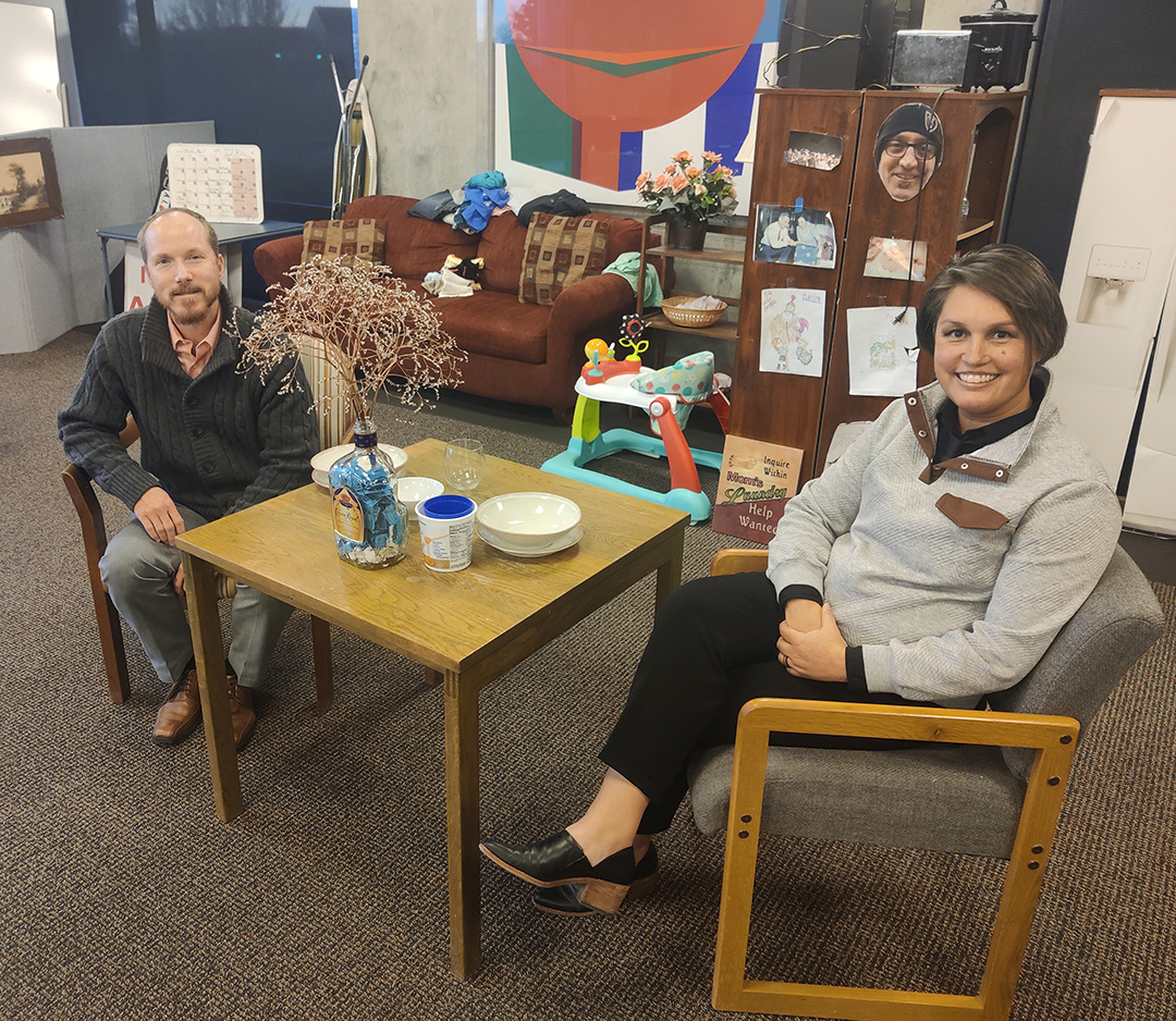 A man and a woman sit at opposite ends of a table and are turned to look at the camera.