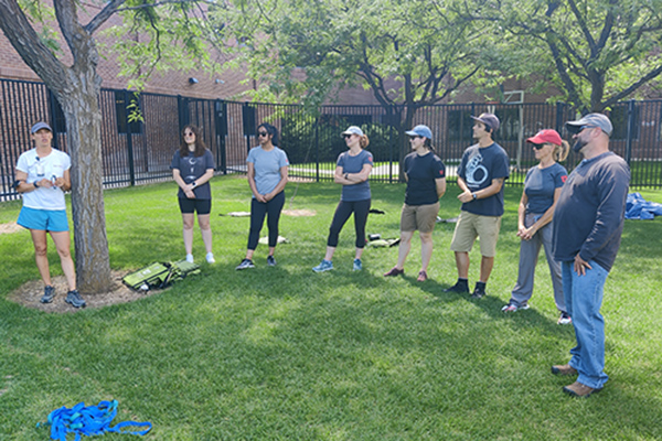 Eight staff stand in a half-circle on grass, with trees in the background.